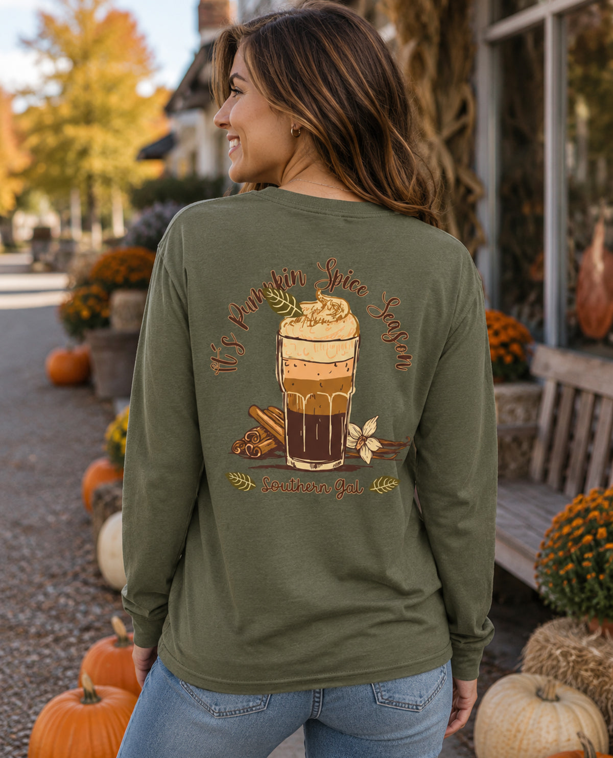 Woman wearing a green long-sleeve shirt with a pumpkin spice latte design in an outdoor setting with pumpkins and fall decorations.