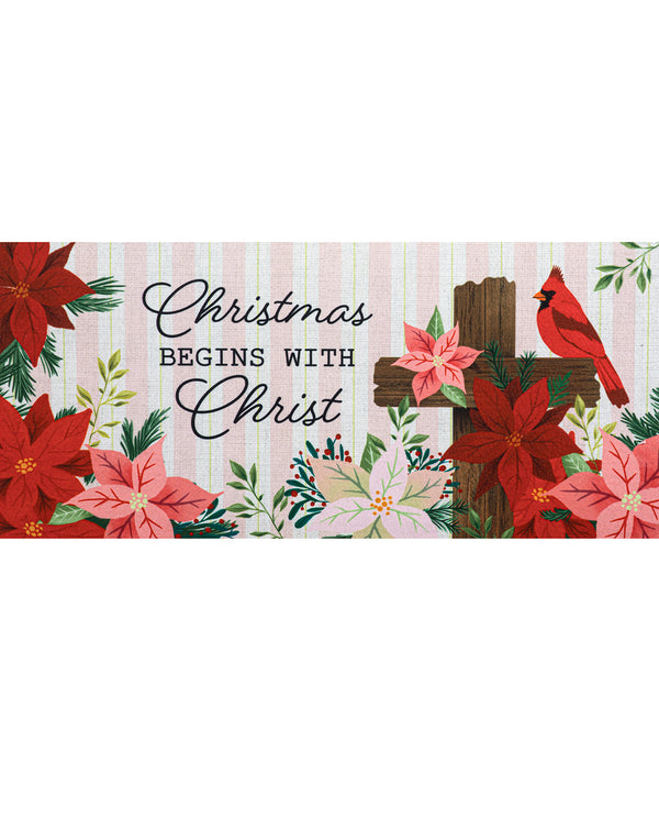 A rectangular Christmas-themed banner or sign with the phrase 'Christmas BEGINS WITH Christ' in black script and serif text. The design features red poinsettias, pink flowers, green foliage, a brown wooden cross, and a red cardinal on a pale pink striped background.