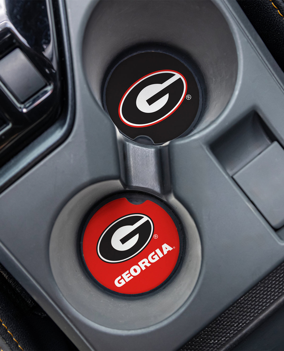 Two circular car cup holder covers with Georgia Bulldogs branding, shown installed in a gray center console. One cover is black with a white and red logo, and the other is red with a black and white logo and the word GEORGIA.