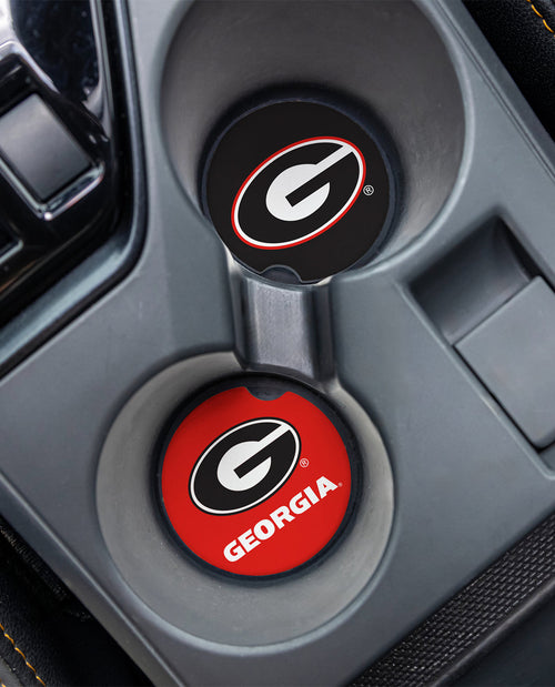 Two circular car cup holder covers with Georgia Bulldogs branding, shown installed in a gray center console. One cover is black with a white and red logo, and the other is red with a black and white logo and the word GEORGIA.