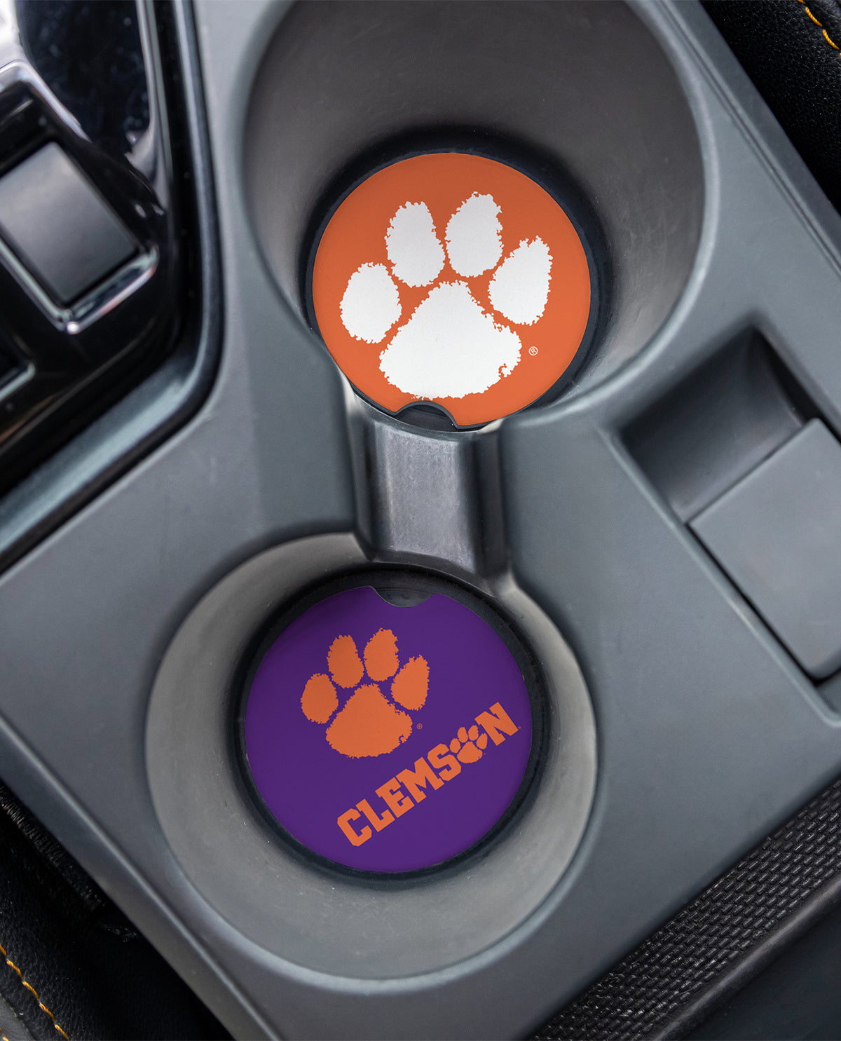 Two Clemson Tigers themed car cup holder coasters shown installed in a gray center console. The top coaster is orange with a white paw print, and the bottom coaster is purple with an orange paw print and the word 'CLEMSON'.
