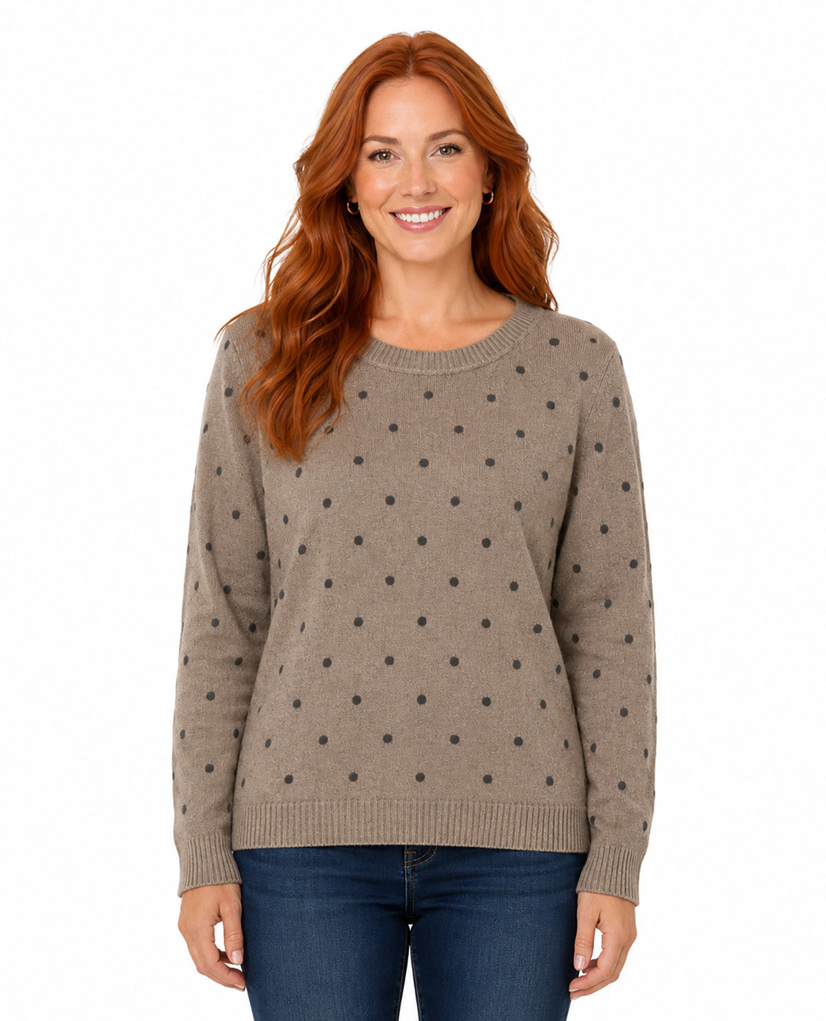 A smiling woman with long red hair wearing a taupe knit sweater with a black polka dot pattern and blue jeans, photographed against a solid white background.