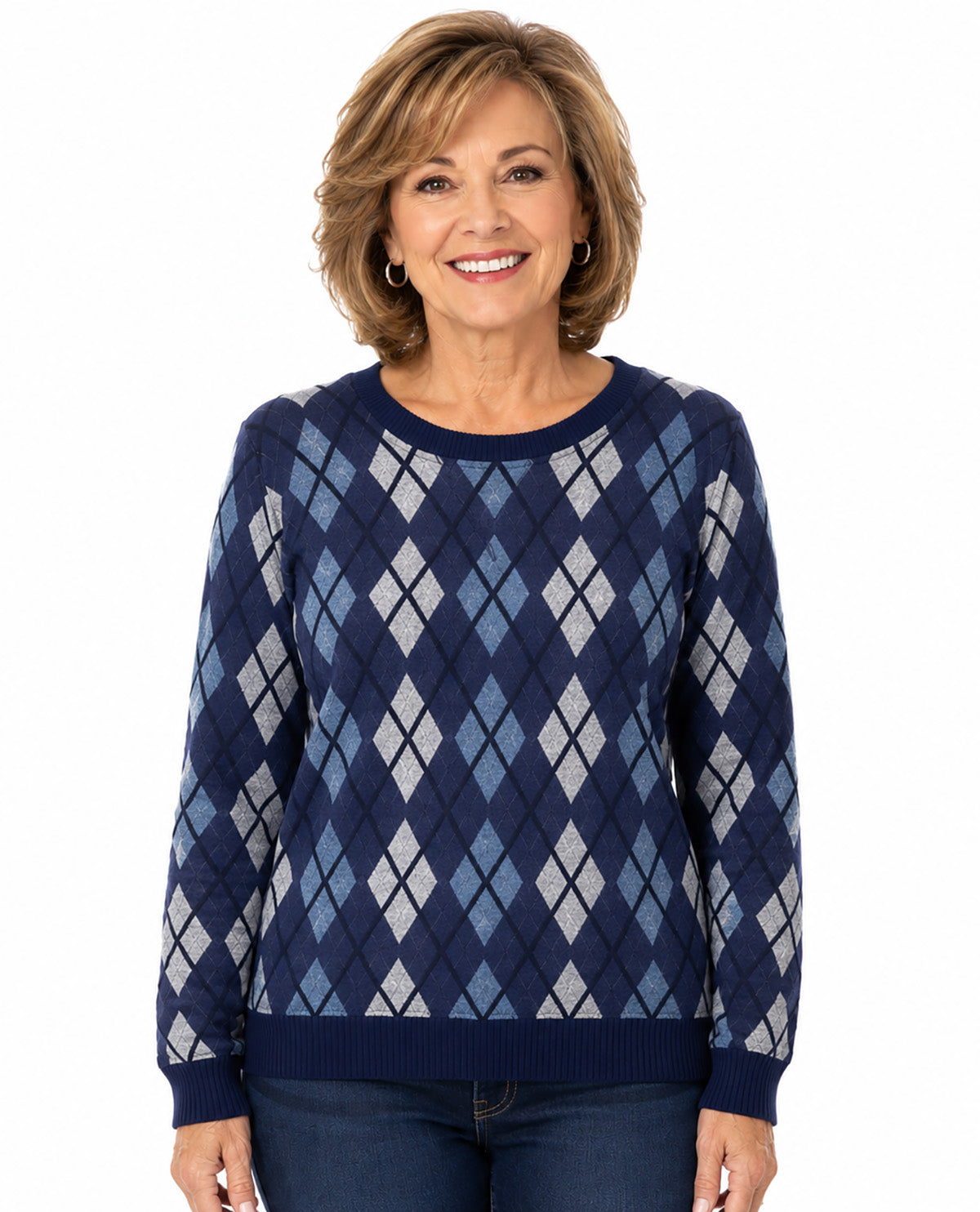 A smiling woman with short brown hair wearing a navy blue long-sleeve sweater with an argyle pattern in navy, light blue, and white, paired with dark blue jeans, against a plain white background.