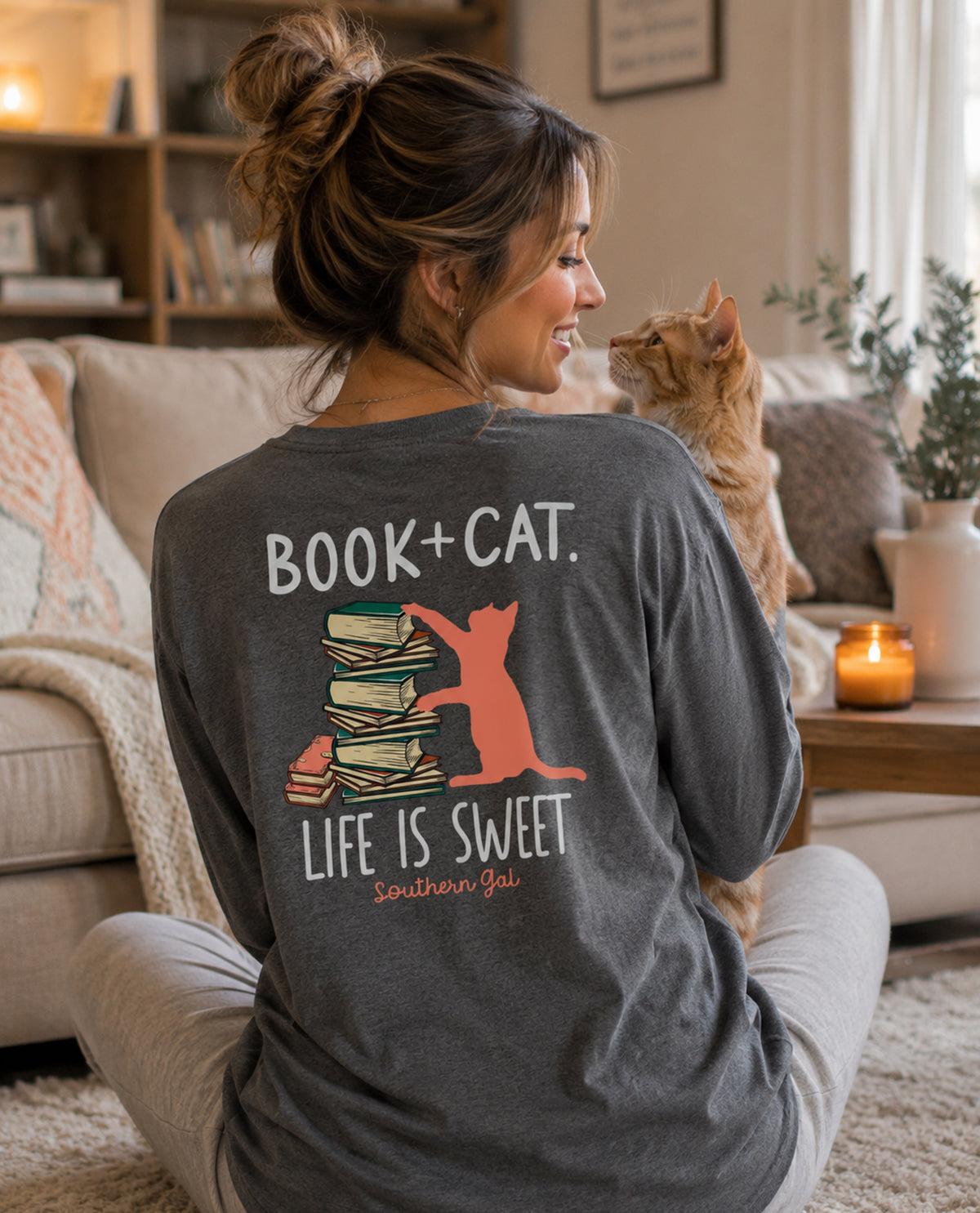Woman wearing a shirt with 'Book + Cat. Life is Sweet' design, sitting with a cat in a cozy living room.