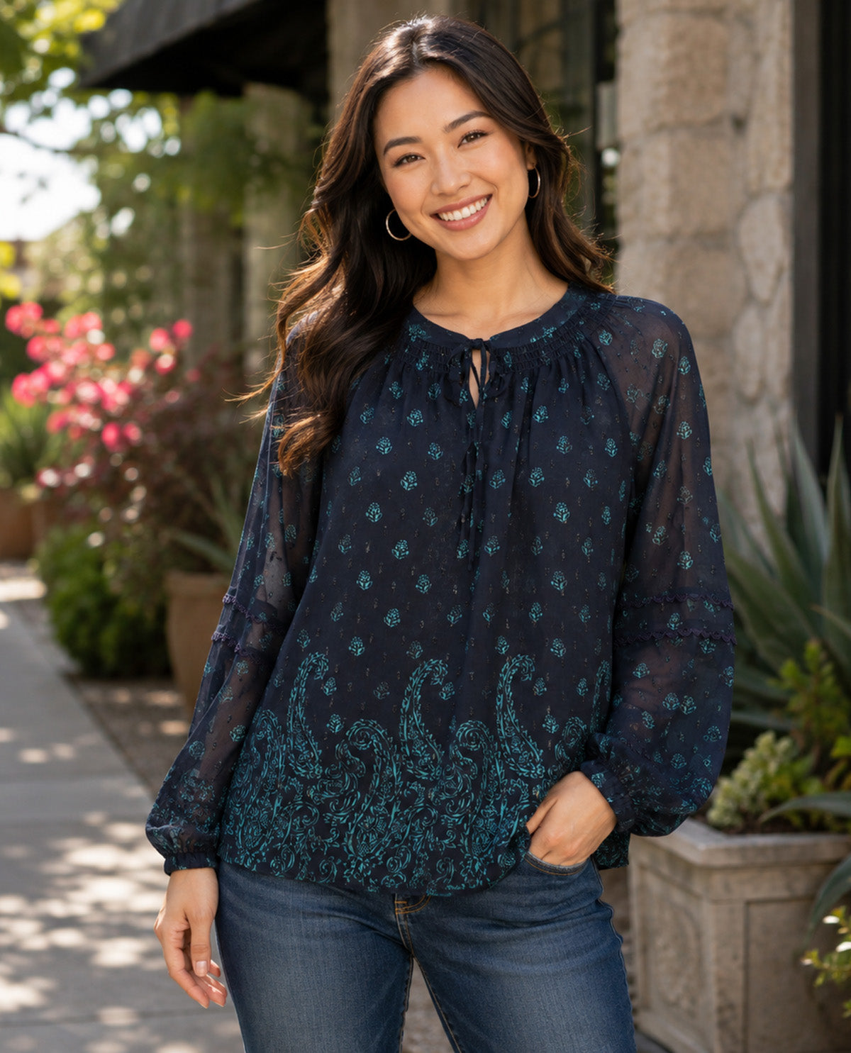 A woman wearing a sheer dark navy long-sleeve blouse with a teal paisley and floral pattern, paired with blue jeans, standing outdoors in front of a stone building.