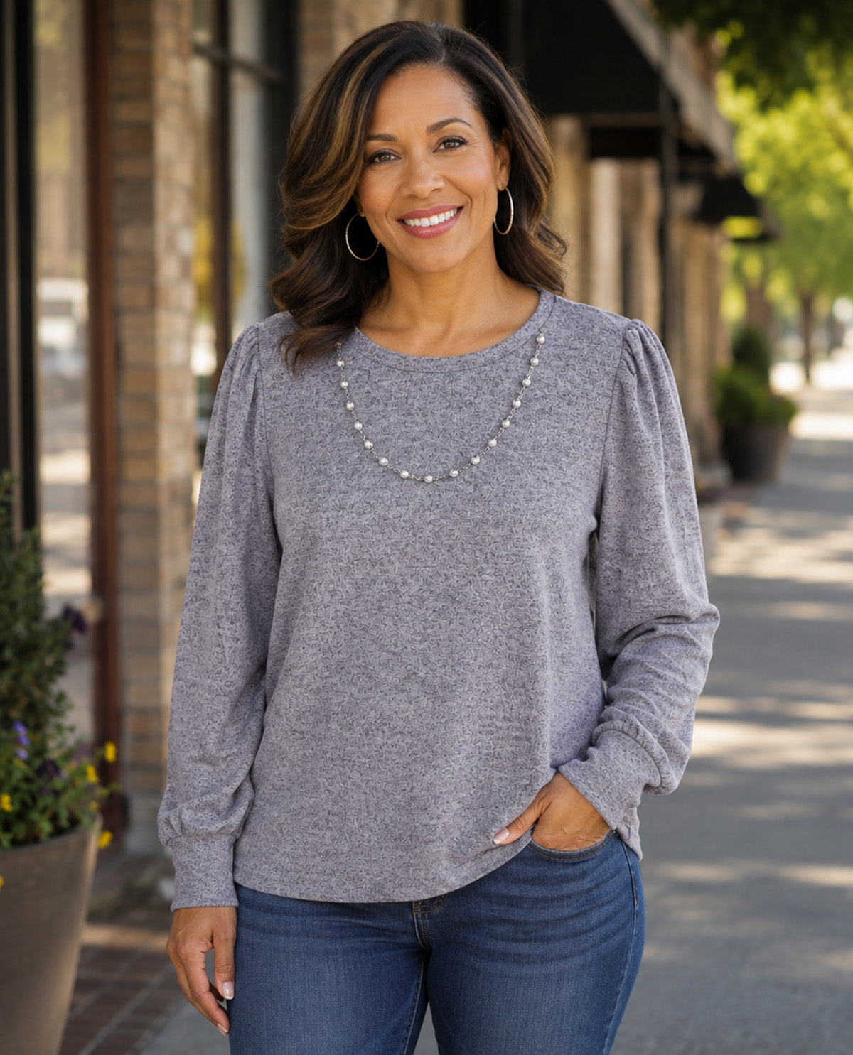 A smiling woman wearing a heather gray long-sleeve top with puffed shoulders and a pearl necklace, standing on a sunny sidewalk in front of storefronts.