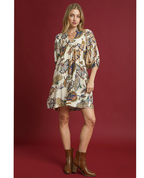 A woman modeling a short, loose-fitting dress with a cream base and an all-over multicolor butterfly and floral print, paired with brown ankle boots, against a solid dark red studio background.