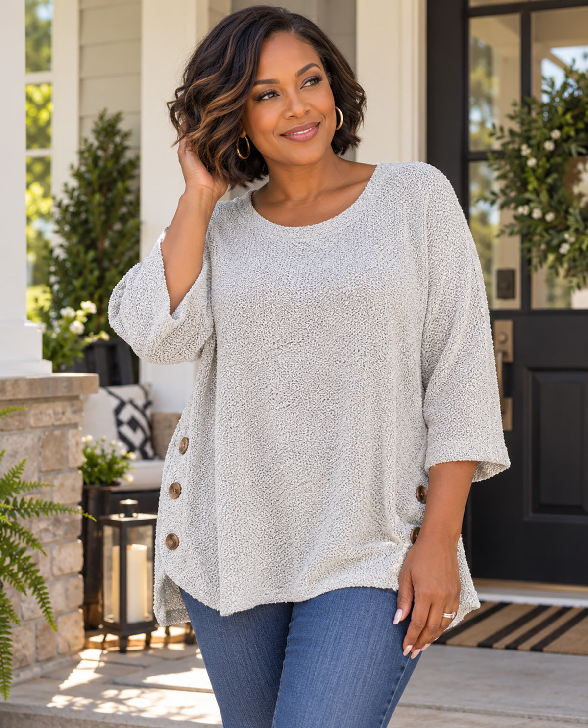 A woman wearing a light beige textured three-quarter sleeve top with brown buttons down the side, paired with blue jeans, standing on a front porch.