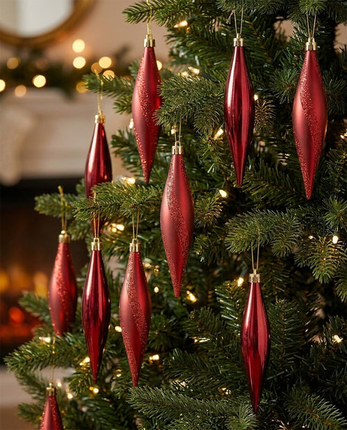 Red teardrop-shaped Christmas tree ornaments with gold caps and subtle glitter, hanging on a green Christmas tree with warm white lights.