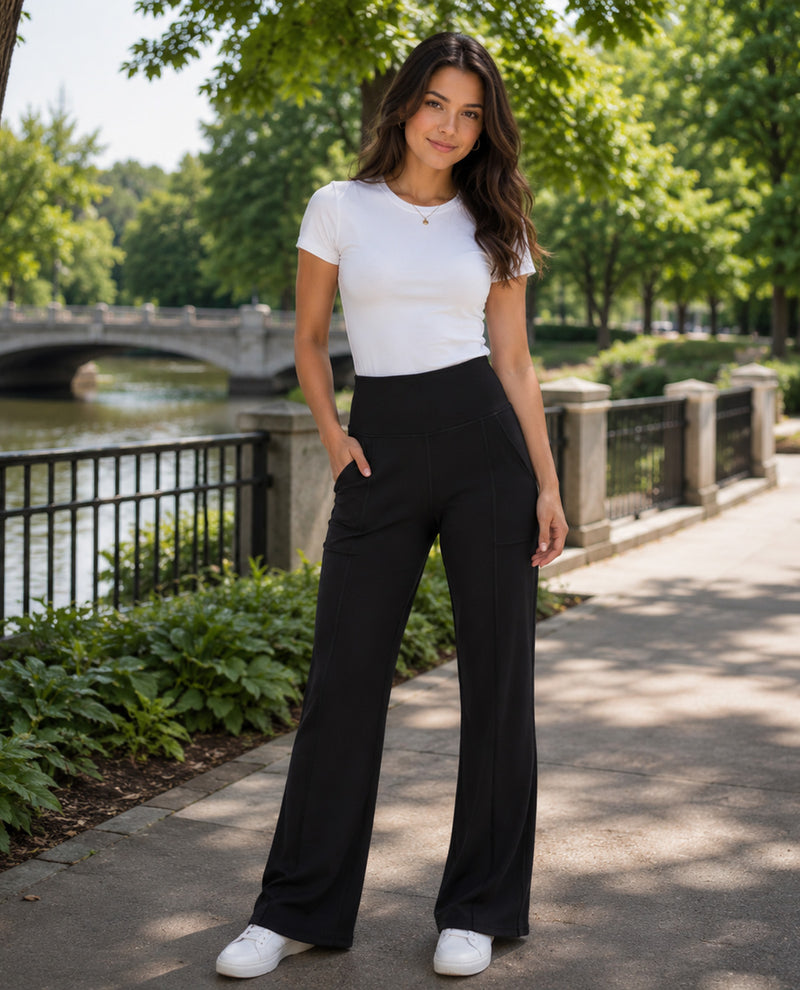 A woman stands outdoors on a paved walkway beside a river, wearing a fitted white short-sleeve T-shirt, black wide-leg pants, and white sneakers. Trees, a bridge, and water are visible in the background.