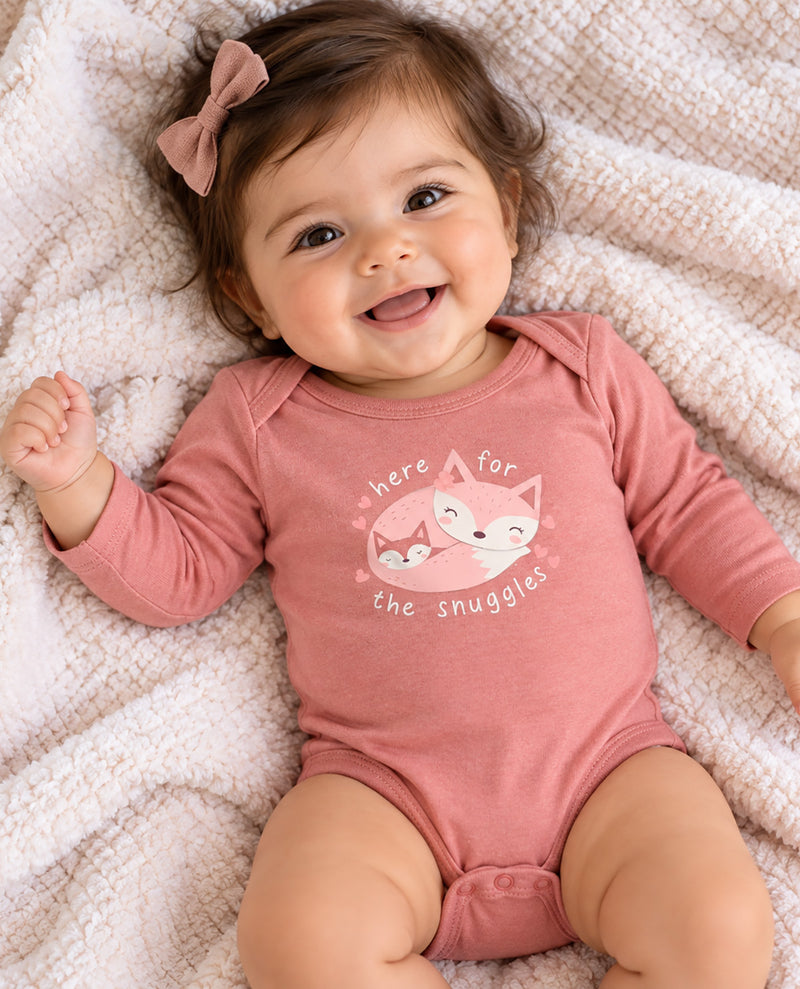A smiling baby lying on a soft cream blanket, wearing a dusty pink long-sleeve bodysuit with a fox graphic and the words 'there for the snuggles.'.