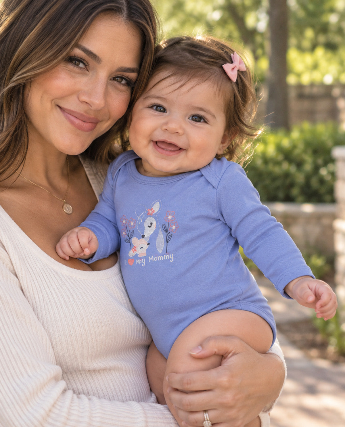 A smiling baby girl wearing a light blue long-sleeve bodysuit with a cartoon bunny and the text 'My Mommy' is held by a woman in a cream ribbed sweater in an outdoor park setting.