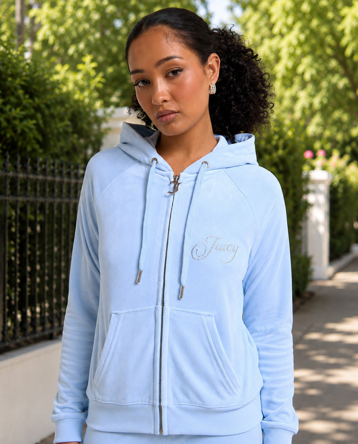 A woman wearing a light blue zip-up hoodie with 'Juicy' embroidered in silver on the chest, standing outdoors on a sunny day with trees and a fence in the background.