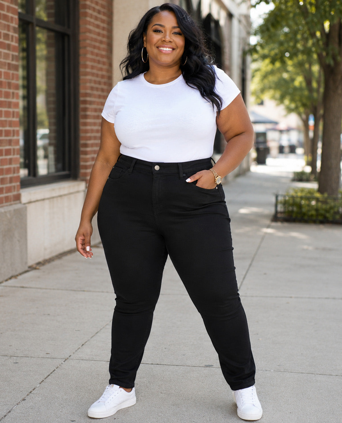 A woman modeling black high-waisted skinny jeans with a white t-shirt and white sneakers, standing on a city sidewalk.