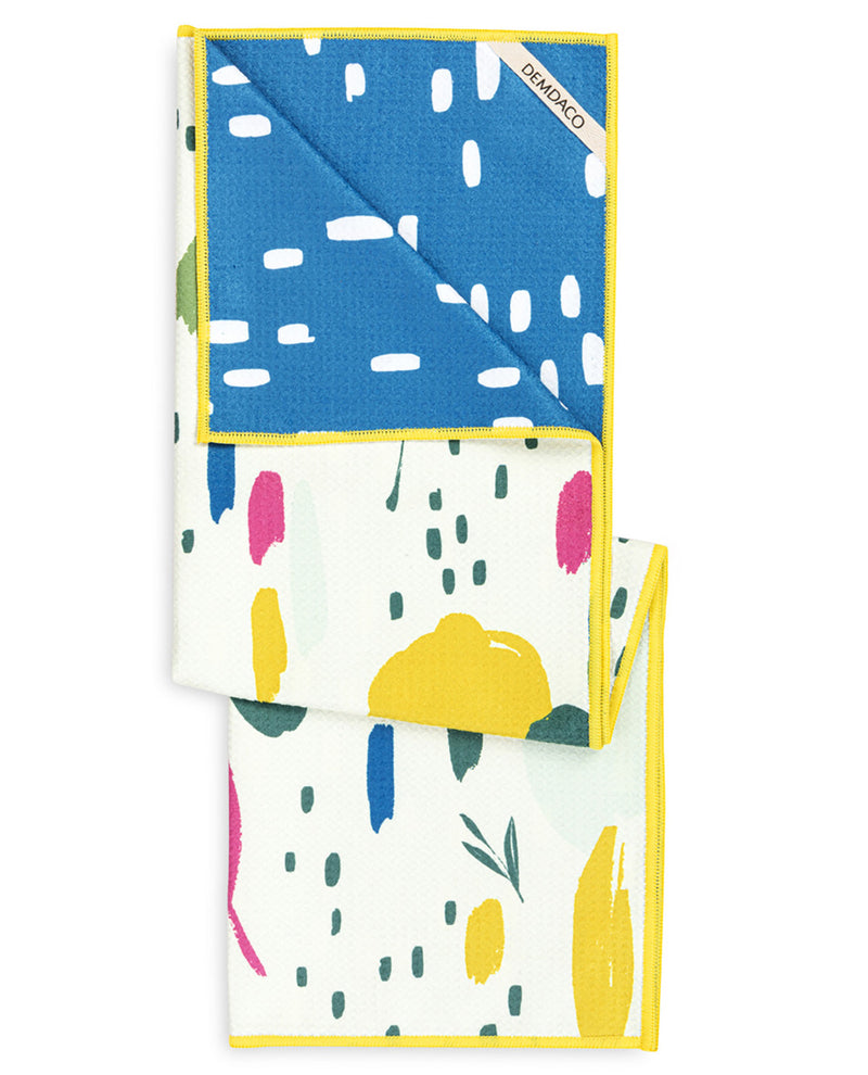 A folded cotton cloth napkin with a colorful abstract print, featuring a blue top section with white brushstrokes and a white bottom section with yellow, pink, blue, and green shapes. It has a bright yellow border and a visible 'DIAMONDO' brand tag.