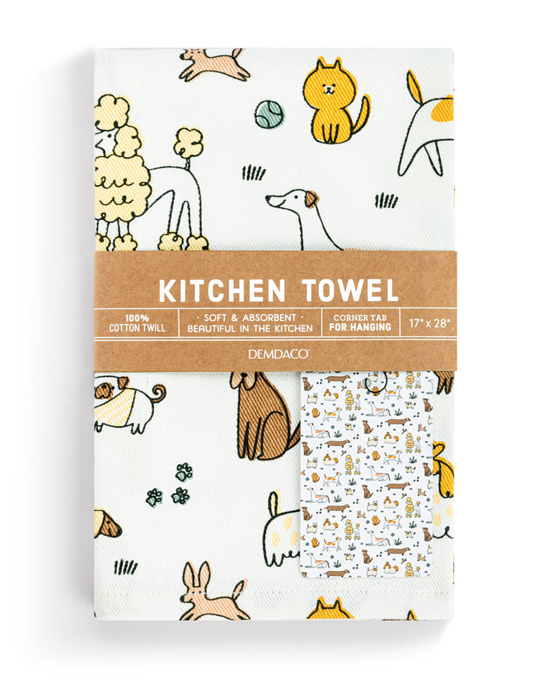 A packaged kitchen towel set by Demdaco with a brown paper band label. The towel features a white background with a playful pattern of various dog breeds in yellow, beige, and brown line drawings.