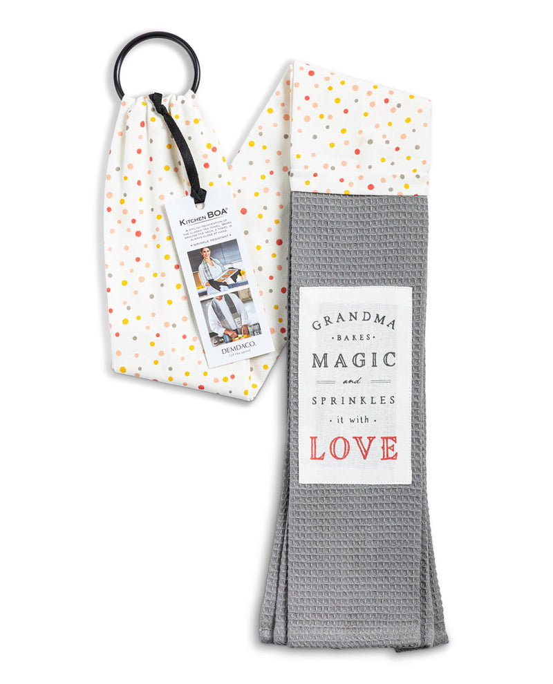 A folded gray waffle-weave kitchen towel with a white label reading 'GRANDMA BAKES MAGIC SPRINKLES it with LOVE', shown next to its white drawstring pouch with multicolored polka dots and a black hanging ring.