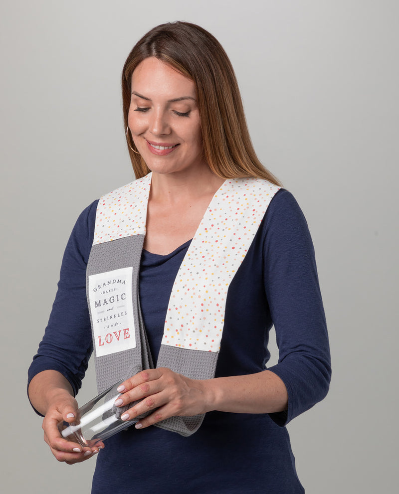 A woman wearing a white and gray fabric vest with colorful polka dots and a printed label that reads 'GREATING THE MAGIC, STRENGTHENING LOVE' holds a small clear glass jar containing white items, against a plain gray studio background.