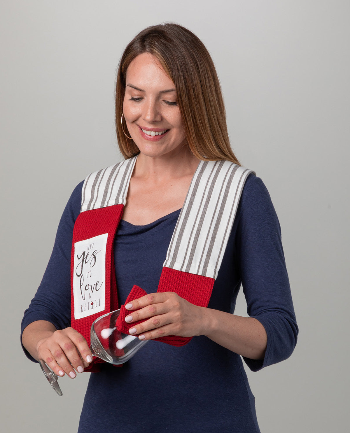 A woman wearing a navy blue long-sleeve shirt models a red and white striped kitchen towel with the phrase 'You Love Me' printed on it. She is holding a wine glass and pouring wine while wearing the towel around her shoulders, set against a plain light gray background.