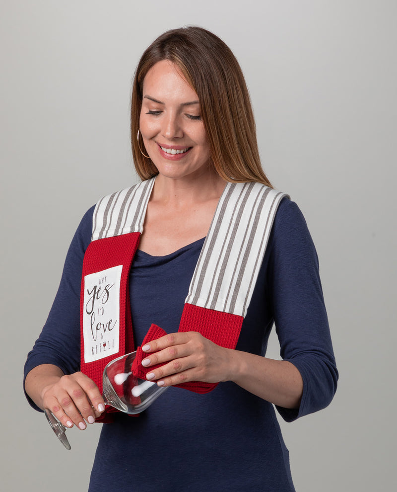 A woman wearing a navy blue long-sleeve shirt models a red and white striped kitchen towel with the phrase 'You Love Me' printed on it. She is holding a wine glass and pouring wine while wearing the towel around her shoulders, set against a plain light gray background.