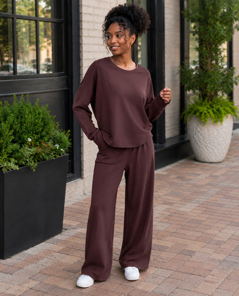 A woman wearing a matching two-piece lounge set in a deep chocolate brown color, standing on a brick walkway in front of a building with potted plants.