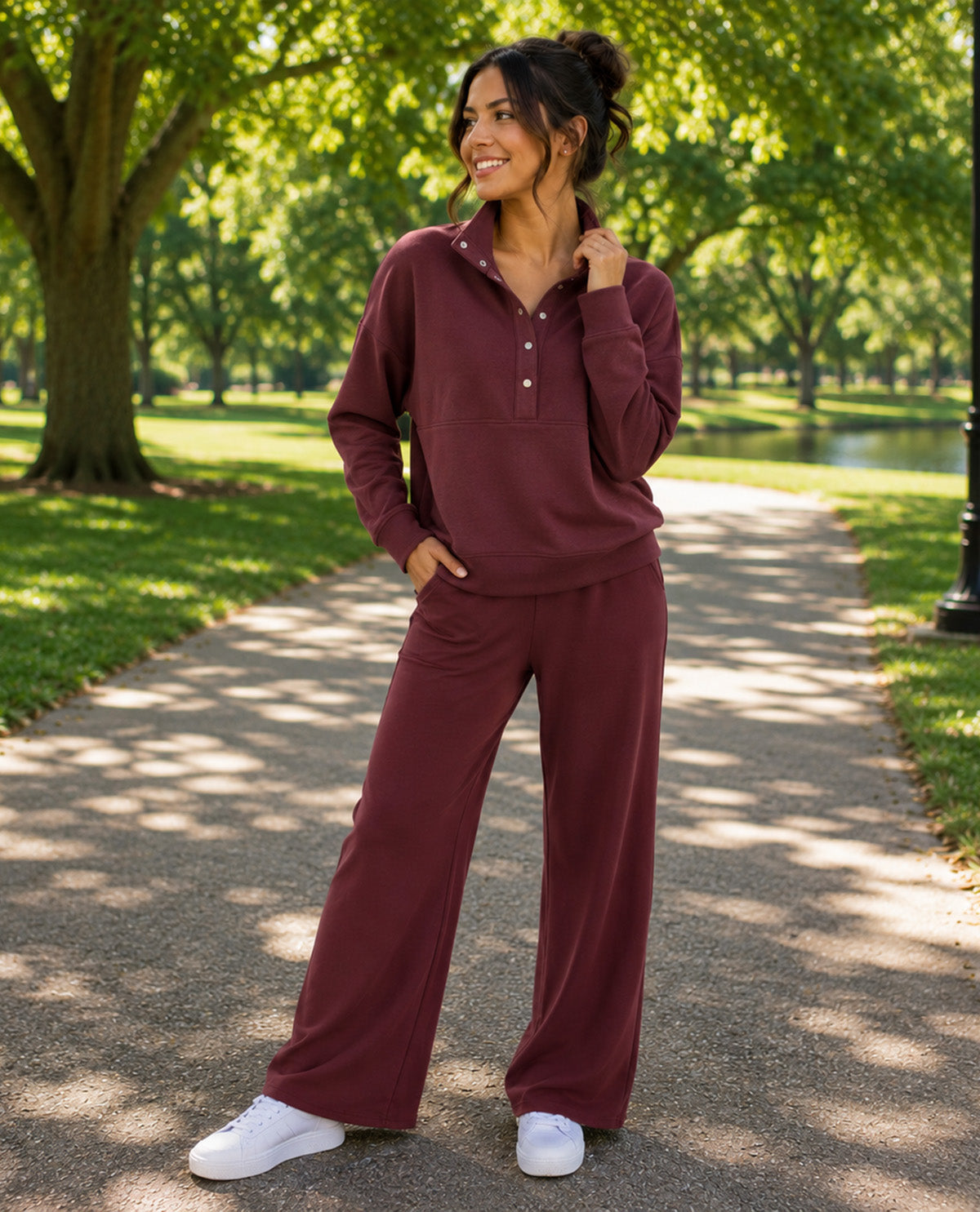 A woman stands on a sun-dappled park path wearing a matching burgundy lounge set consisting of a long-sleeve polo-style top with a button placket and wide-leg pants, paired with white sneakers.