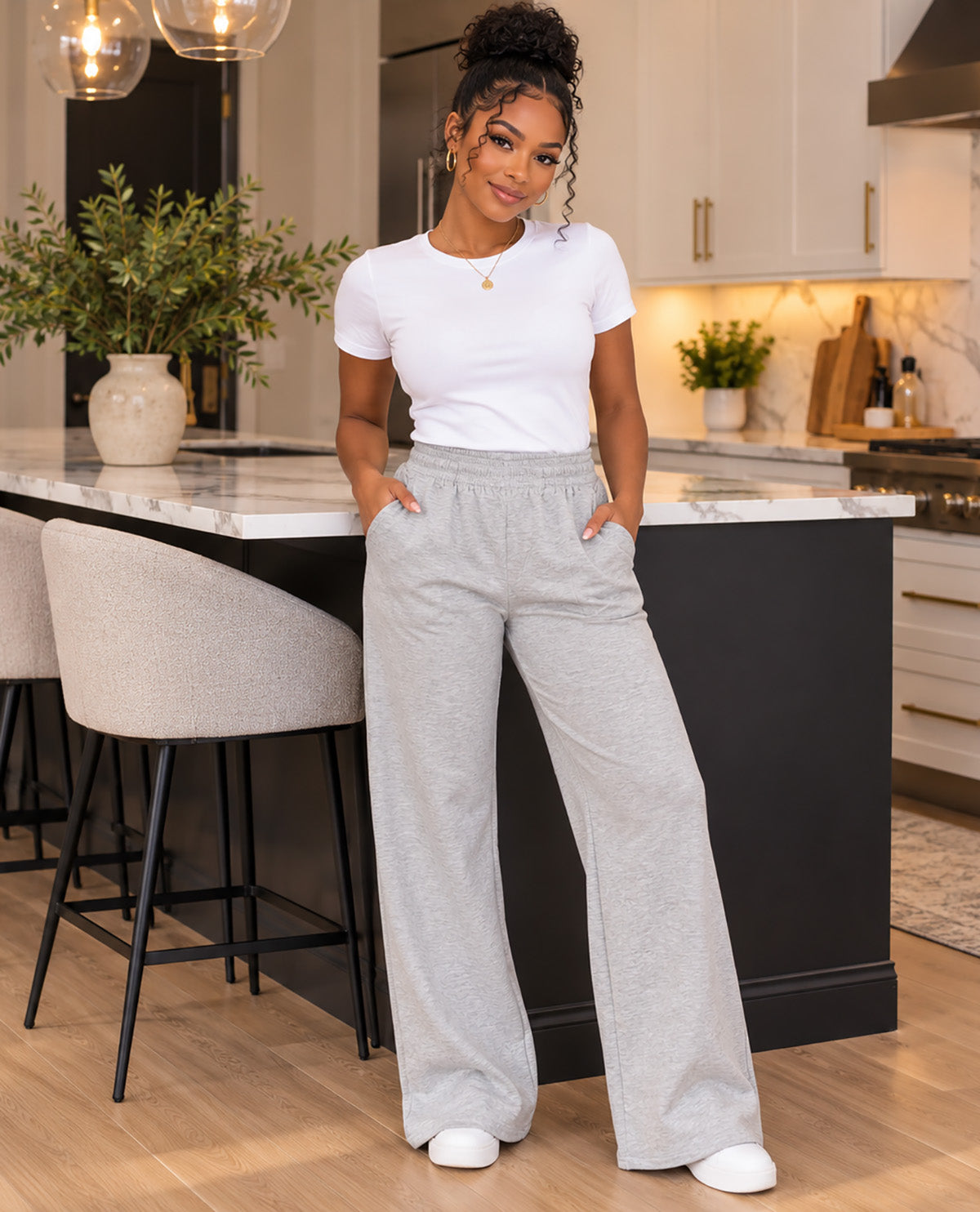 A woman stands in a modern kitchen wearing a white short-sleeve crewneck t-shirt and light gray wide-leg sweatpants. She is positioned in front of a kitchen island with bar stools.