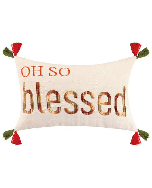 An off-white rectangular throw pillow featuring the phrase 'OH SO blessed' in orange and brown plaid lettering, with red tassels at each corner and green fabric accents.