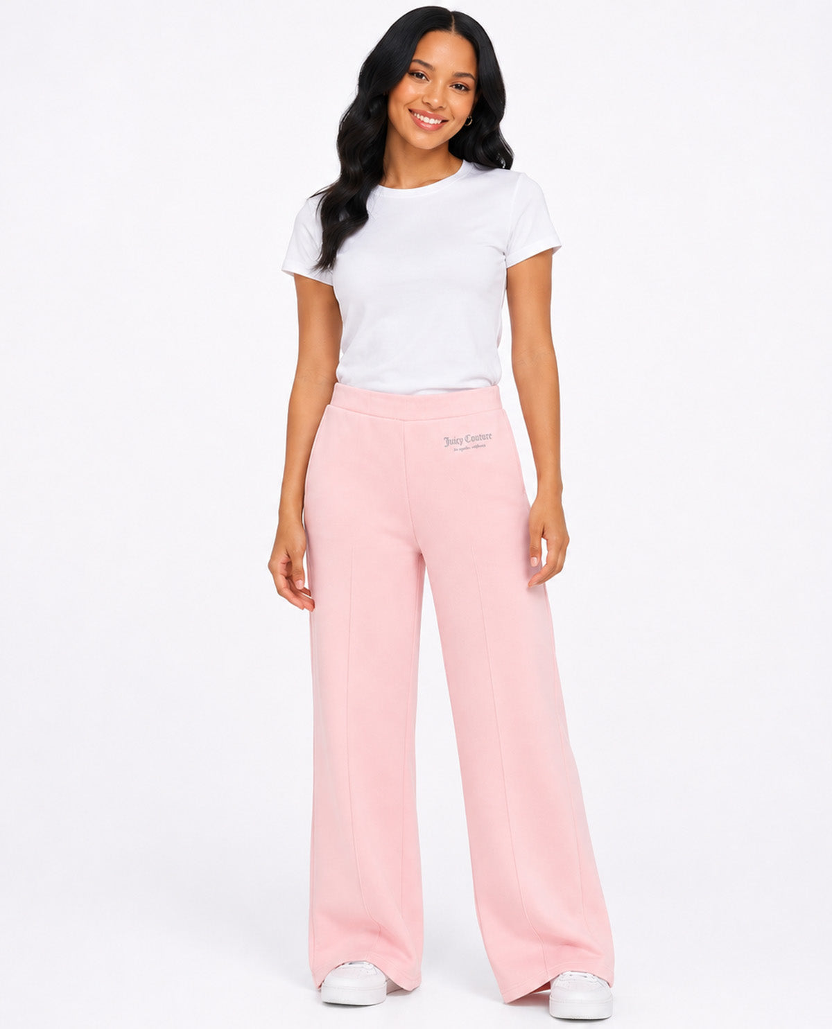 A woman modeling a pair of light pink wide-leg sweatpants with a small embroidered logo on the left hip, paired with a white t-shirt and white sneakers, against a plain white studio background.
