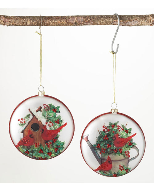 Two round hanging Christmas ornaments displayed on a tree branch against a white background. Each ornament features a winter garden scene with red cardinals, green holly, red berries, a birdhouse, and a watering can.