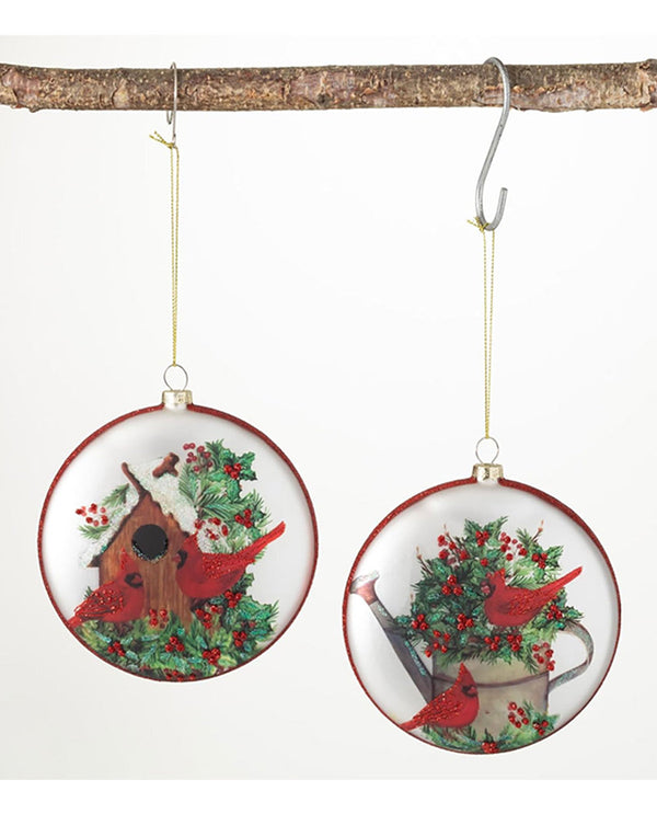 Two round hanging Christmas ornaments displayed on a tree branch against a white background. Each ornament features a winter garden scene with red cardinals, green holly, red berries, a birdhouse, and a watering can.