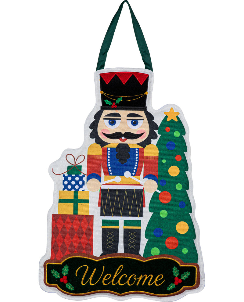 A festive Christmas nutcracker wall hanging with a green hanging loop, featuring a colorful nutcracker, a decorated Christmas tree, wrapped gifts, and a black banner at the bottom that reads 'Welcome' in gold script.