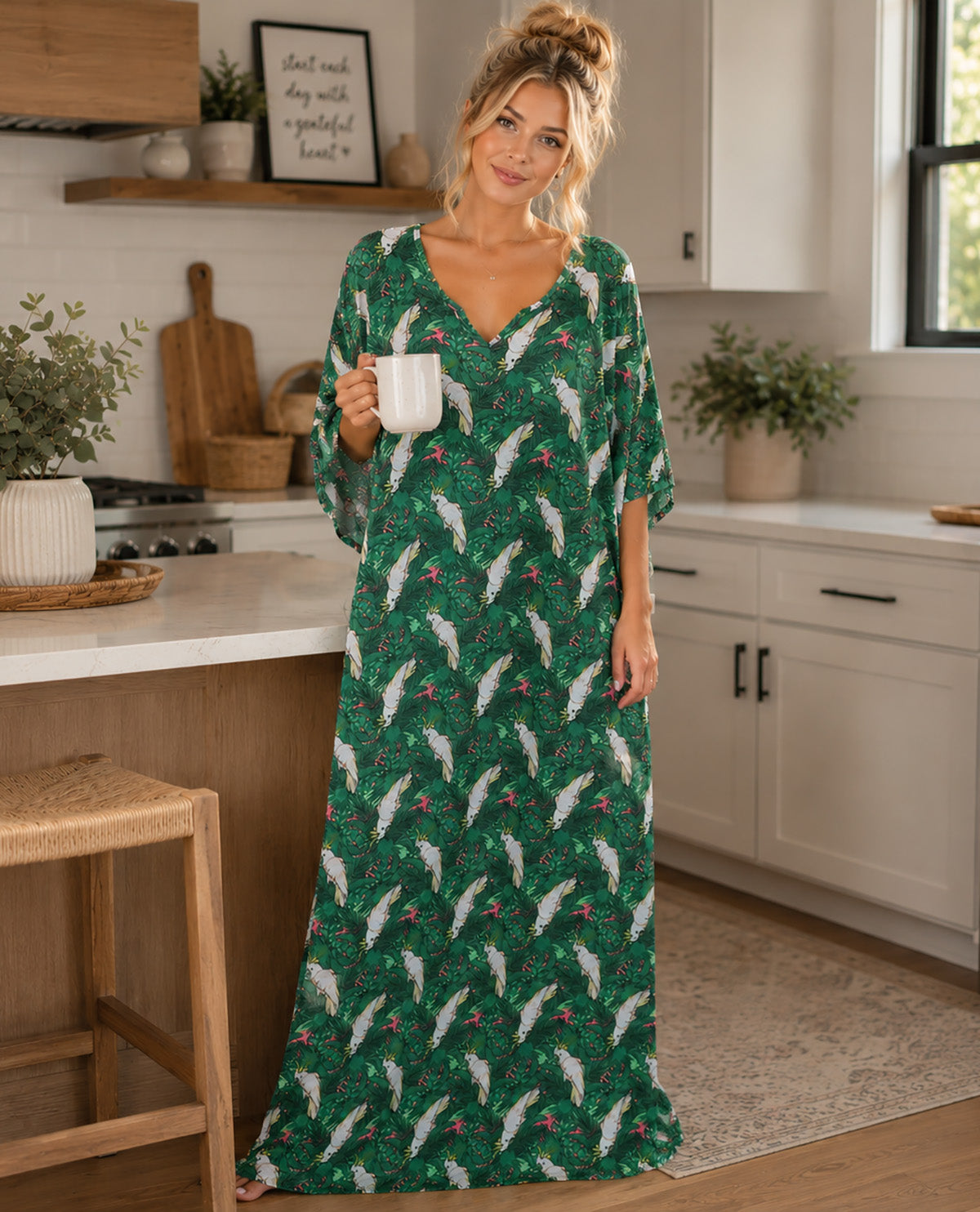A woman stands in a bright kitchen wearing a long green maxi dress with a white and red holiday pattern, holding a white mug.