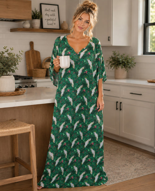 A woman stands in a bright kitchen wearing a long green maxi dress with a white and red holiday pattern, holding a white mug.