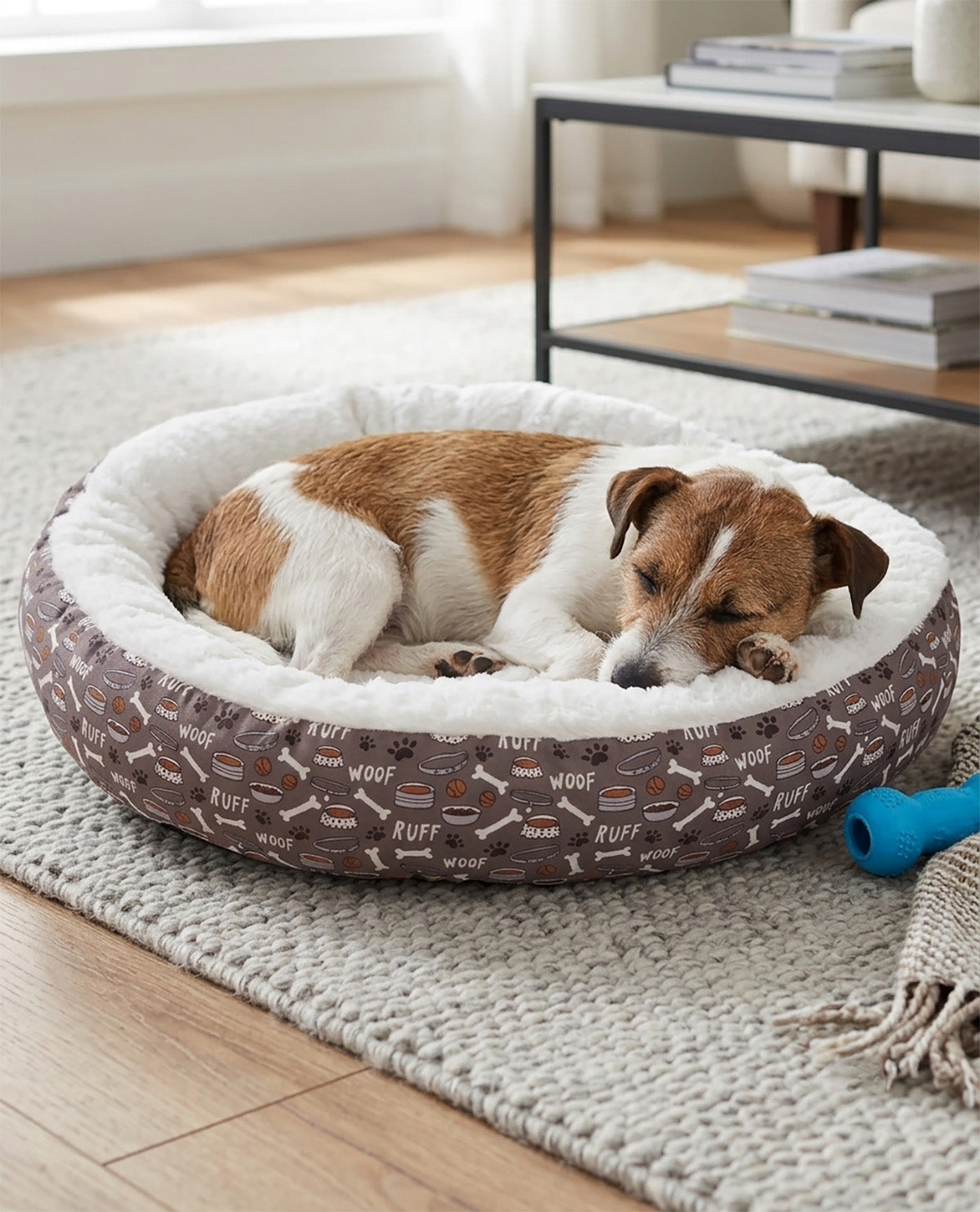 A brown and white dog sleeping comfortably inside a round pet bed with a brown patterned exterior and a white sherpa lining, placed on a light-colored rug in a bright room.