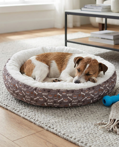 A brown and white dog sleeping comfortably inside a round pet bed with a brown patterned exterior and a white sherpa lining, placed on a light-colored rug in a bright room.