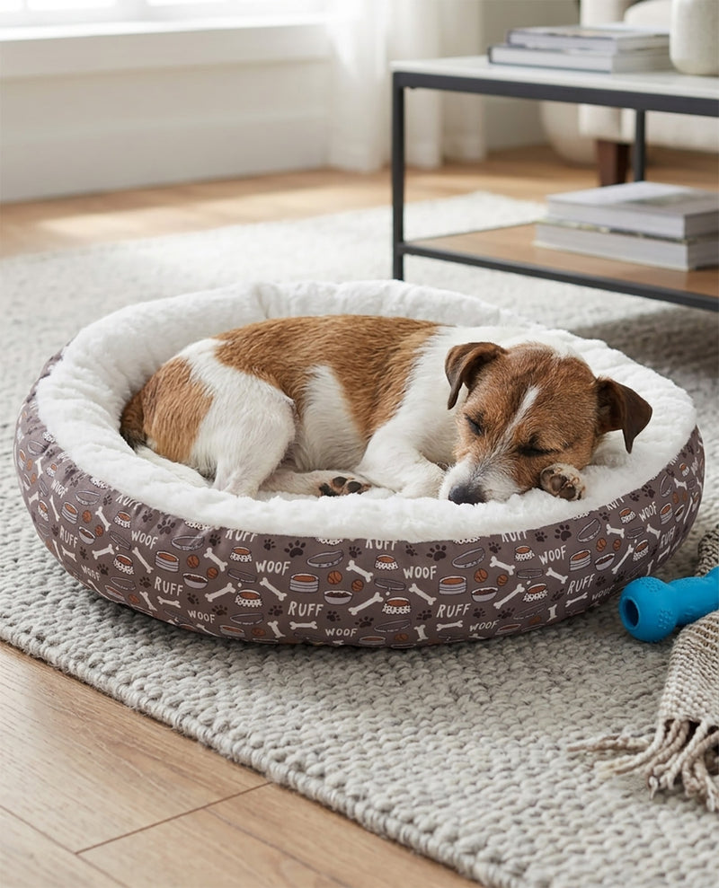 A brown and white dog sleeping comfortably inside a round pet bed with a brown patterned exterior and a white sherpa lining, placed on a light-colored rug in a bright room.
