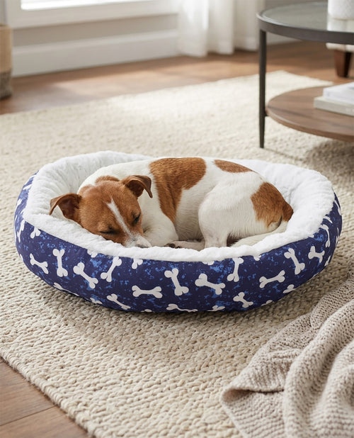A small brown and white dog sleeping comfortably inside a round navy blue dog bed with white bone patterns and a white plush lining, placed on a beige woven rug in a cozy room.