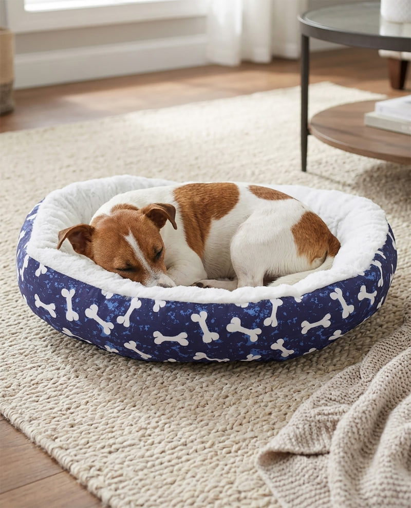 A small brown and white dog sleeping comfortably inside a round navy blue dog bed with white bone patterns and a white plush lining, placed on a beige woven rug in a cozy room.