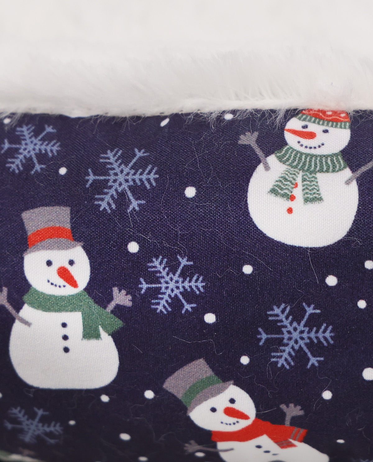 Close-up of a fabric surface with a navy background printed with white snowflakes and snowmen wearing red, green, and gray holiday accessories, with a white fuzzy trim at the top.