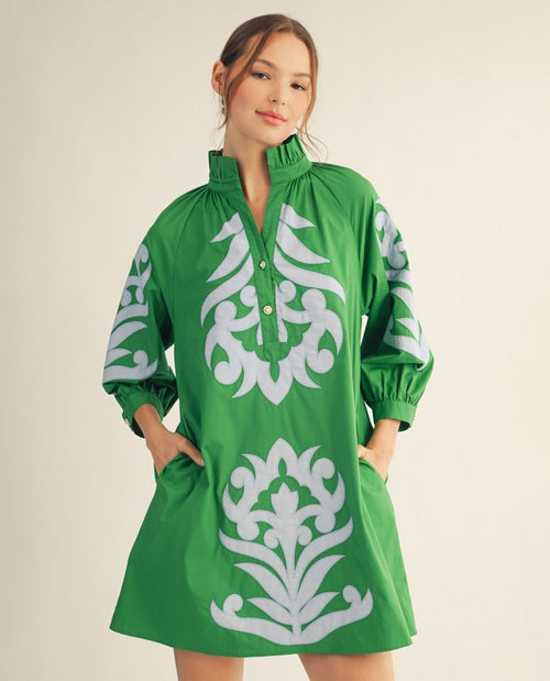 A woman modeling a bright green tunic dress with large white ornamental floral motifs on the front and sleeves, photographed against a plain light beige studio background.