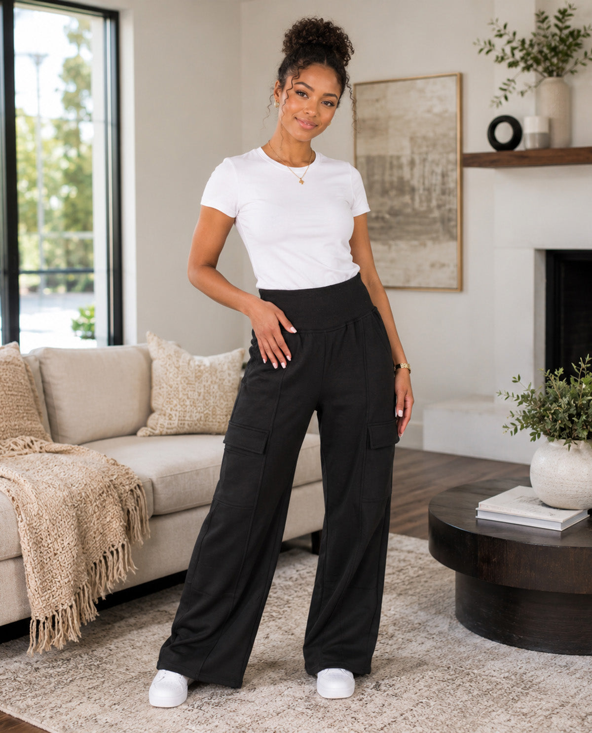 A woman modeling black wide-leg cargo pants with a white t-shirt in a modern living room setting.