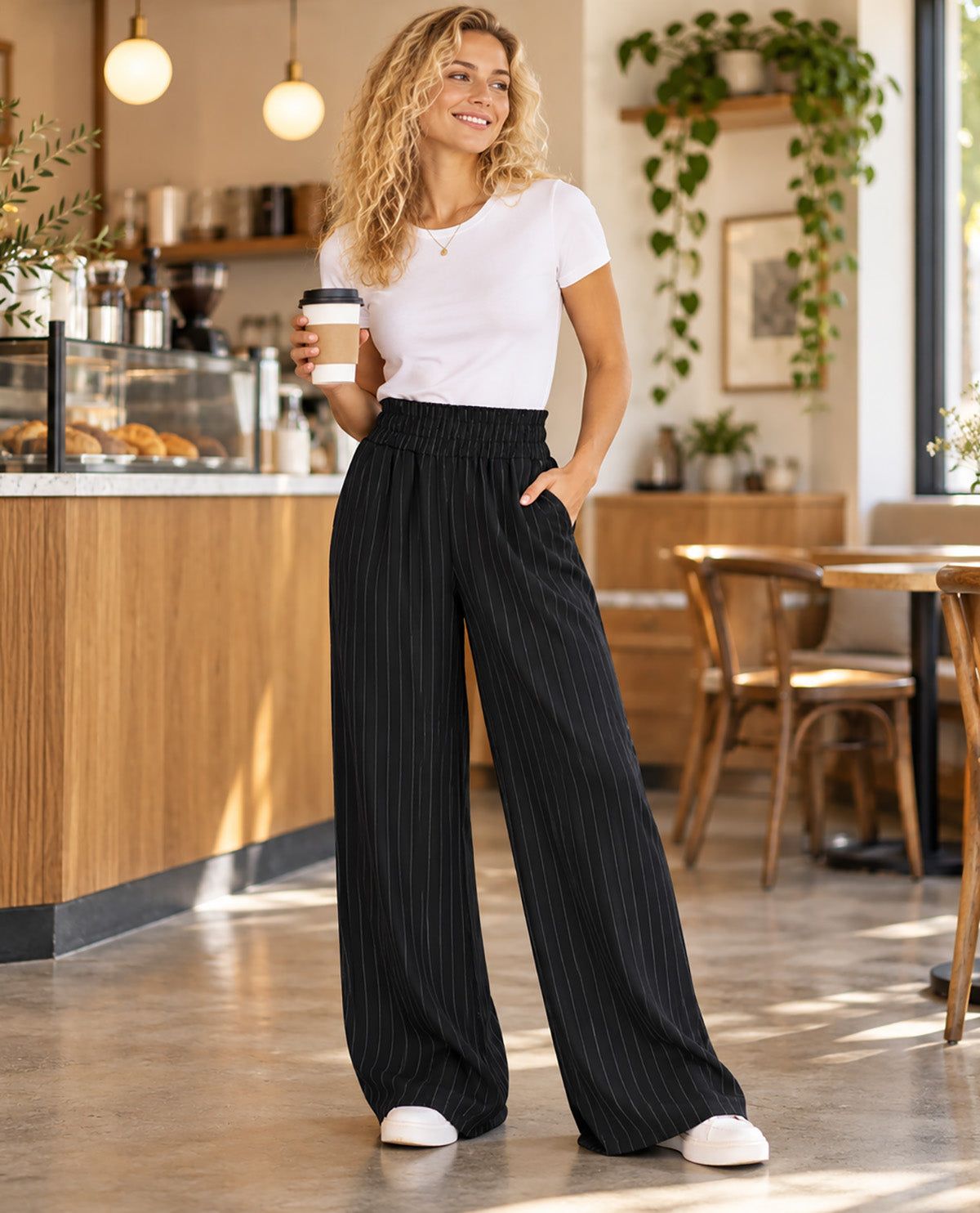 A woman standing in a cafe wearing a white t-shirt and black pinstripe wide-leg pants, holding a coffee cup.