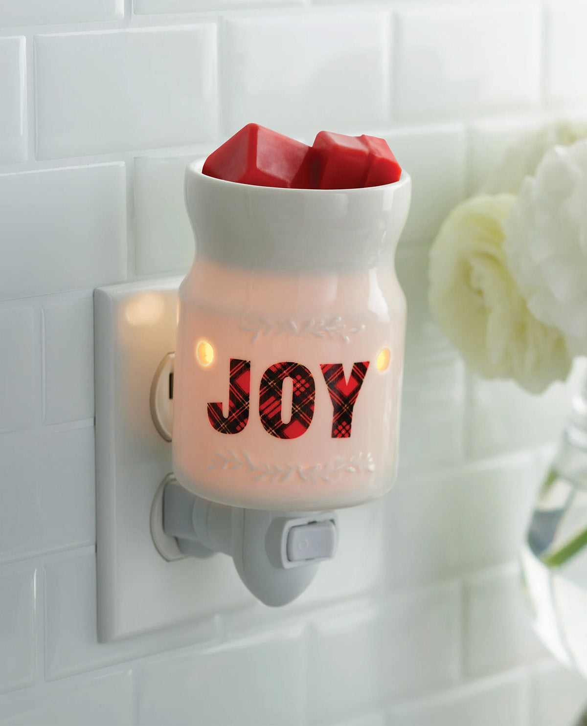 A white ceramic holiday night light plug-in with the word 'JOY' in red plaid letters, glowing with warm light, set against a white subway tile wall.