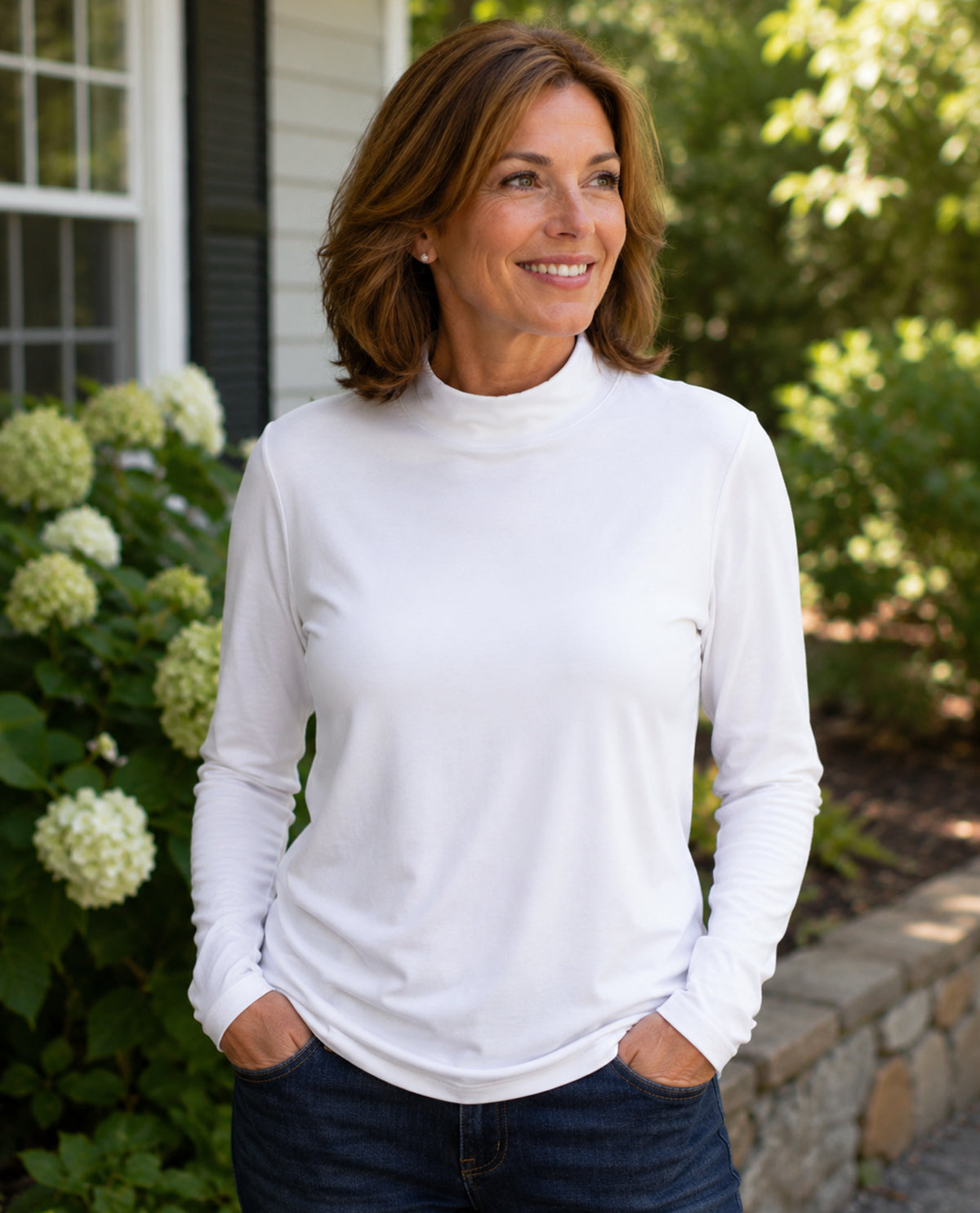 Woman wearing a white long-sleeve top standing outdoors with greenery in the background