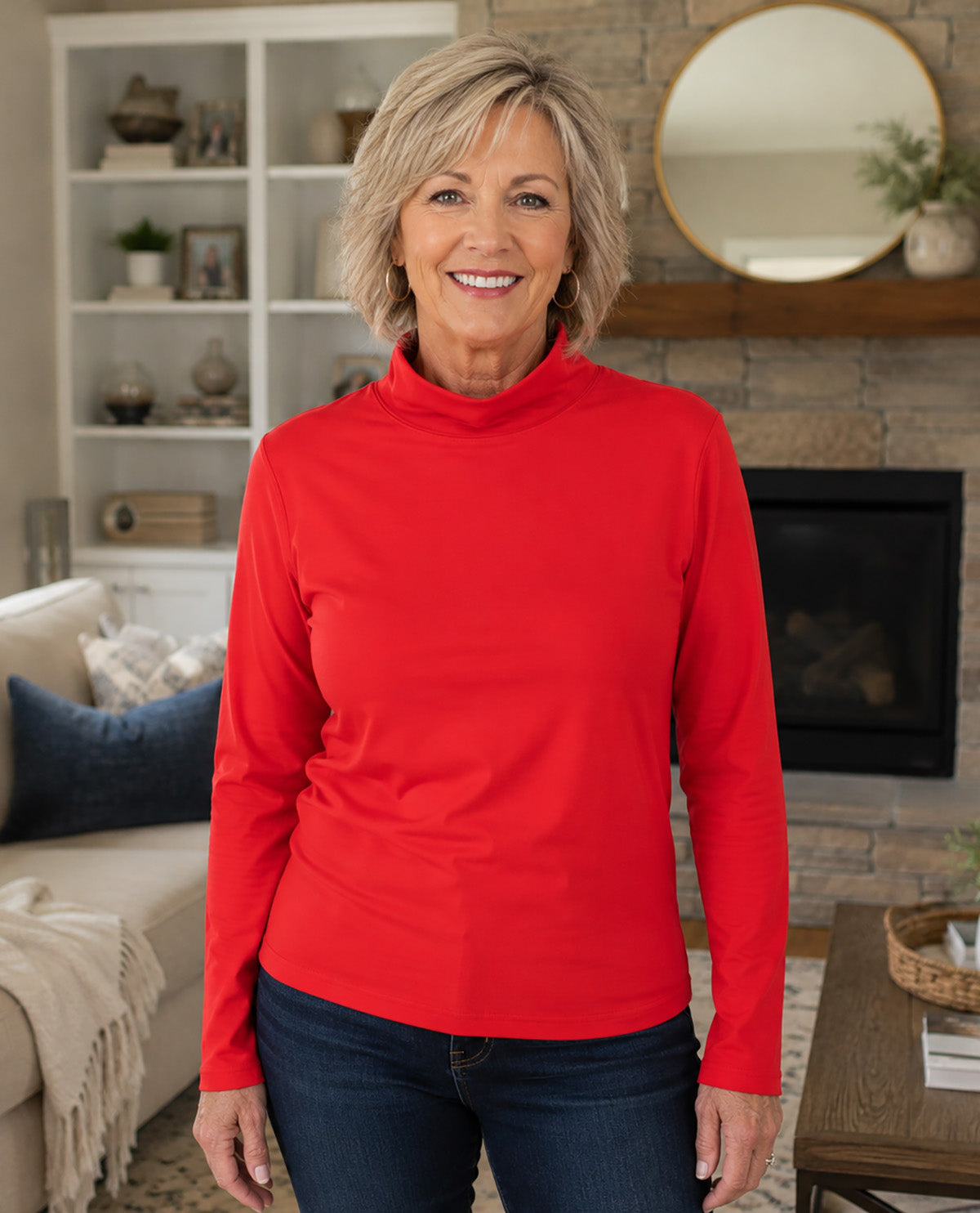 A smiling woman with blonde hair wearing a bright red long-sleeve mock neck top and dark blue jeans, standing in a cozy living room with a fireplace and stone wall background.