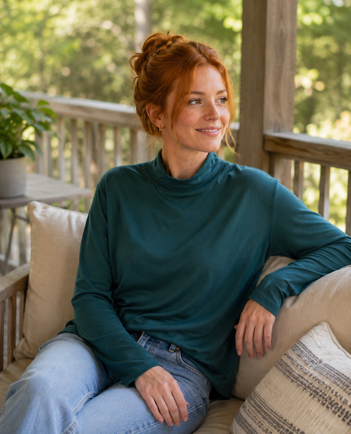 Woman sitting on a porch wearing a teal sweater and blue jeans, surrounded by greenery.