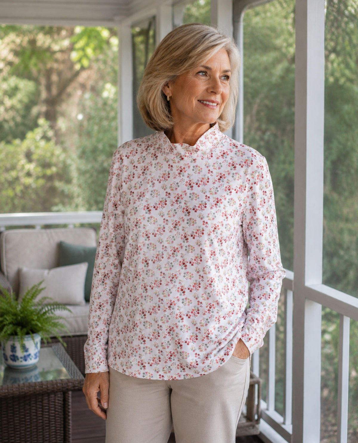 A woman with blonde hair stands on a porch wearing a long-sleeve high-neck blouse with a small red and blue floral print on a white background, paired with beige pants.