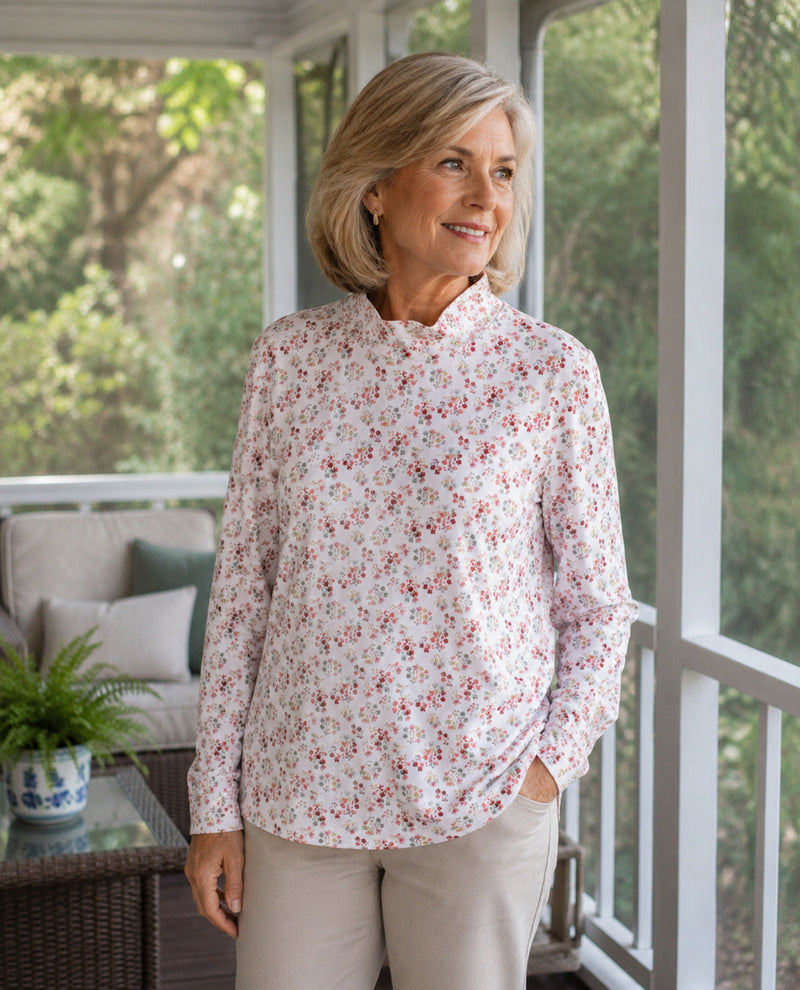 A woman with blonde hair stands on a porch wearing a long-sleeve high-neck blouse with a small red and blue floral print on a white background, paired with beige pants.
