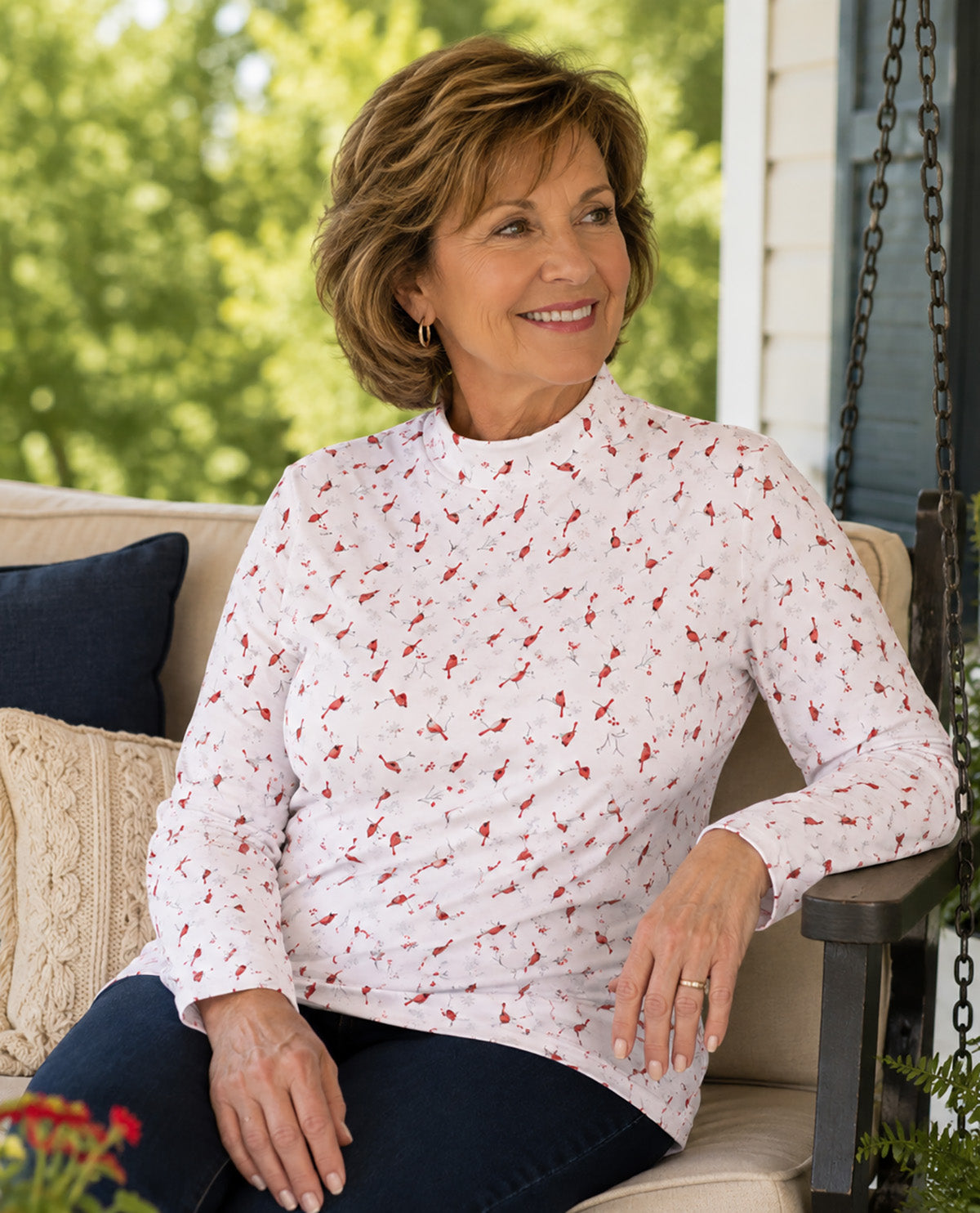 A smiling woman seated on a porch chair wearing a white long-sleeve mock-neck top with a small red allover floral print, paired with dark blue jeans. The background shows a softly blurred outdoor porch setting with green foliage and neutral cushions.